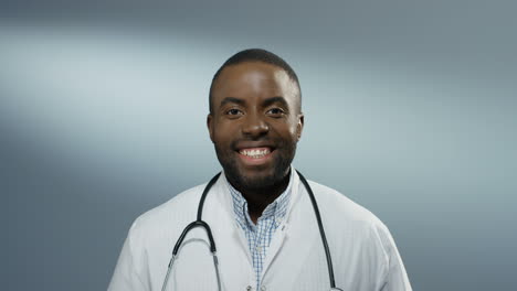 Close-up-of-the-African-American-young-happy-male-doctor-or-intern-smiling-cheerfully-to-the-camera-on-the-gray-wall-background.-Portrait.